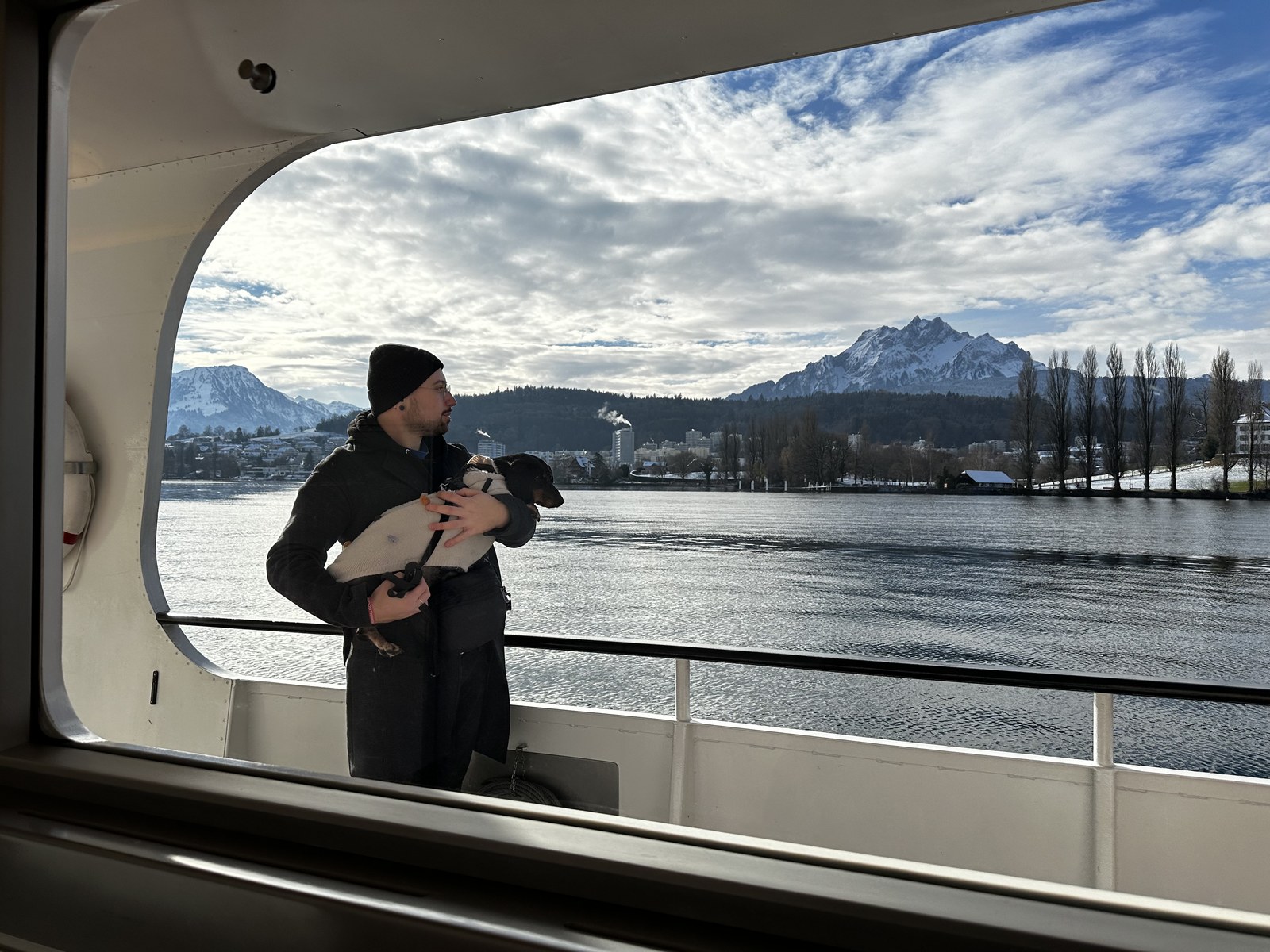 Seen through the window of a boat cabin. A man in a dark coat and black beanie stands on the open deck holding a black and tan dachshund wrapped in a cream knitted jumper. Behind them the lake, the snow covered Pilatus and a winter shoreline.