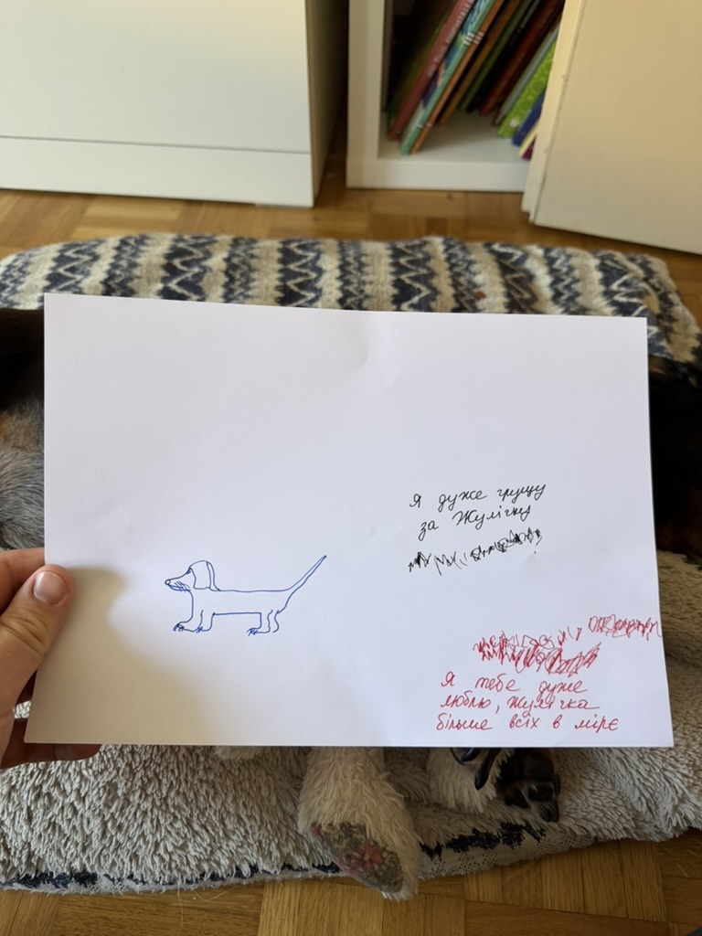 A sheet of white paper held up in one hand. On the left, a line drawing of a dachshund in blue pen. On the right, two lines of adult handwriting in blue and red with a child's red scribbles across and beneath them.